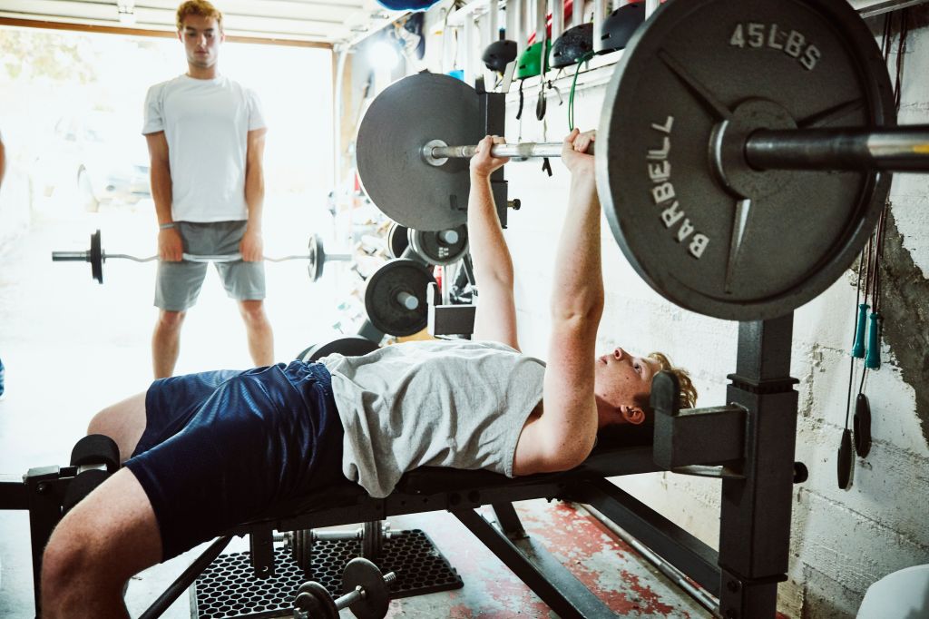 Young man doing bench presses while working out with friends in gym in garage