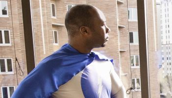 A black businessman office super hero stands at an office window and ponders his next business move.