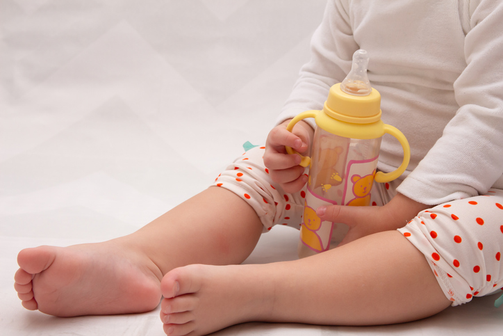 Low Section Of Baby Holding Milk Bottle On Bed At Home
