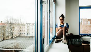 Young casual businesswoman with smart phone sitting on window sill
