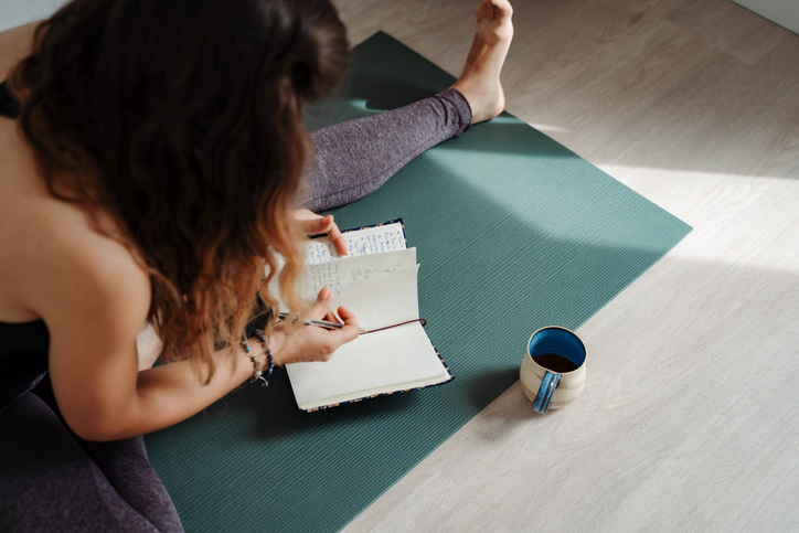 Woman sitting on a yoga mat and writing in a notebook journal and drinking tea coffee from a mug