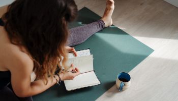 Woman sitting on a yoga mat and writing in a notebook journal and drinking tea coffee from a mug