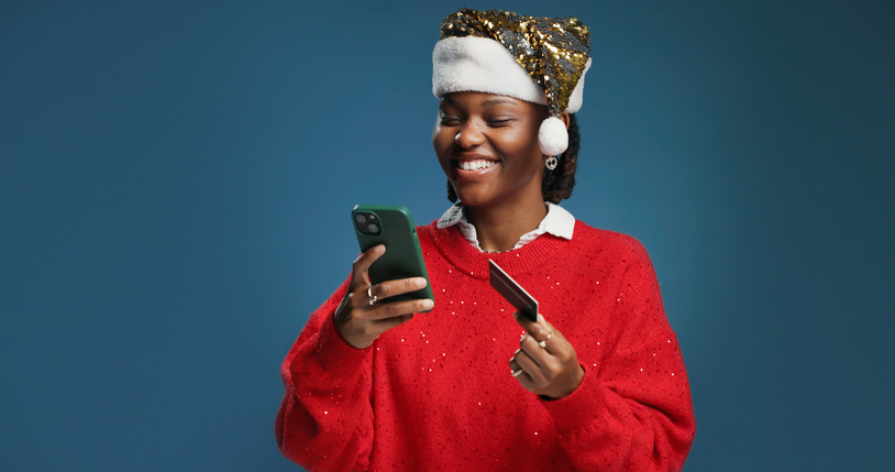 Happy, black woman and credit card with phone for christmas shopping on a blue studio background. Female person, online shopper or mobile smartphone for app, festive purchase or ecommerce on space