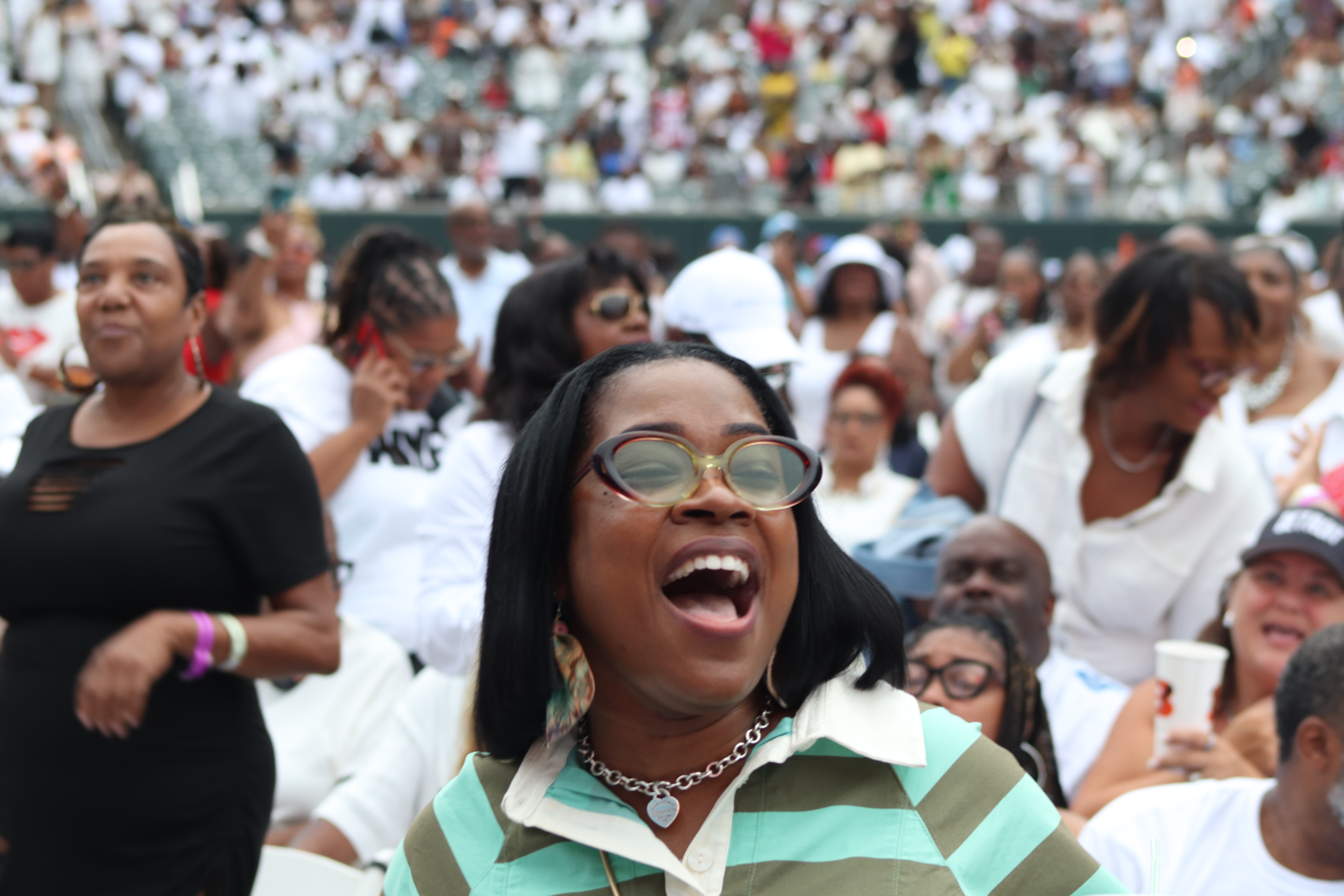 A large crowd of people gathered at an event, with a woman in the foreground laughing and wearing sunglasses.