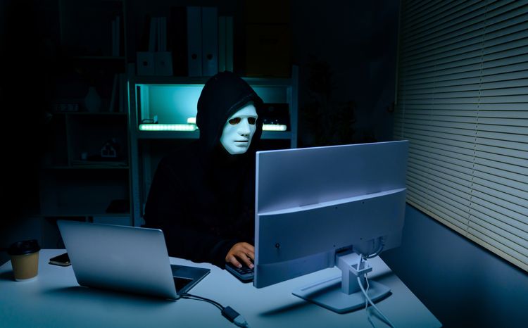 Anonymous hacker wearing mask sitting at desk with laptop and monitor in dark room. cybersecurity concept for it security campaigns, data breach prevention, and technology blogs.